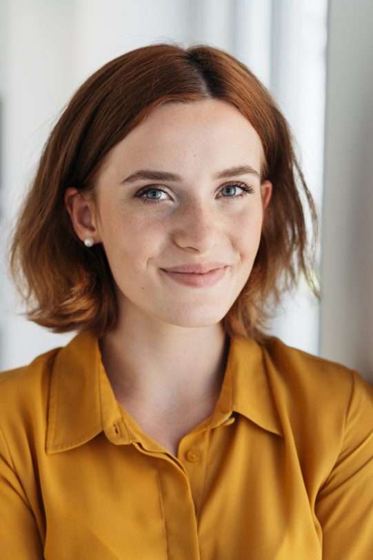 Attractive young businesswoman with a happy smile leaning against an office wall looking at the camera in a close up portrait